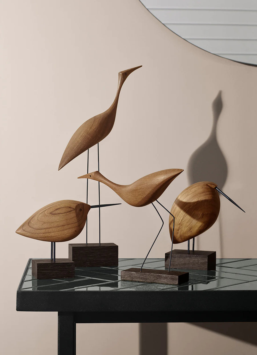 Four Warm Nordic Beak Bird Tall Heron Oak figurines, are elegantly displayed on a table in front of a mirror. The minimalist Danish design perfectly showcases the beauty of natural wood.