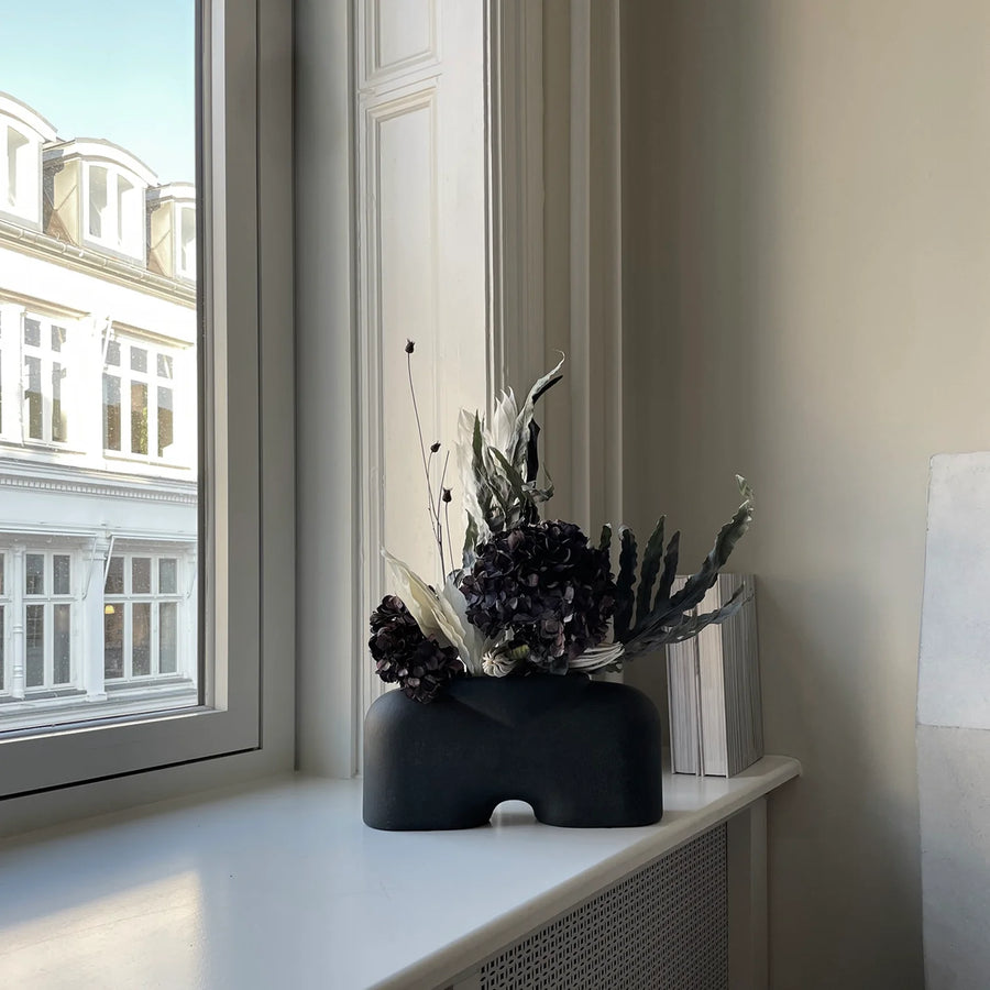 A black vase, known as the 101Cph Cobra Fat Medio Black 212004, gracefully sits on a window sill.