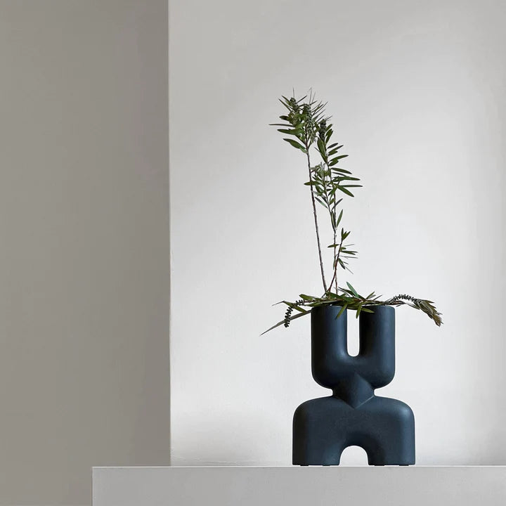 A quirky 101Cph Cobra Double Mini Black 203025 vase from 101 Copenhagen with a plant in it, sitting on top of a white wall.