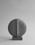 A 101Cph Guggenheim Mini Dark Grey 203005 concrete vase on a grey background, inspired by the Cobra Arts Movement, by 101 Copenhagen.